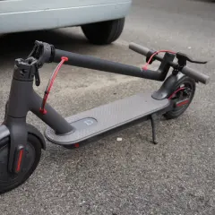 Trottinette électrique grise, avec sa potence repliée, garée sur le bitume à côté d'une voiture.