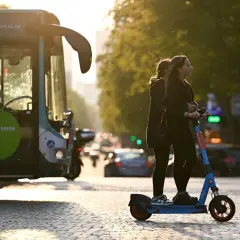 Deux jeunes femmes attendent sur une trottinette électrique, baignées par la lumière du soleil couchant. Un bus RATP est garé à gauche.