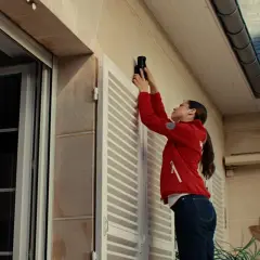 Une femme installe un appareil, peut-être une xiaomi alarme, sur le mur extérieur d'une maison, près d'une fenêtre.