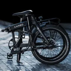 Un vélo électrique carbone pliant, noir, sur un pavé mouillé. Son design moderne et épuré promet des trajets urbains fluides.
