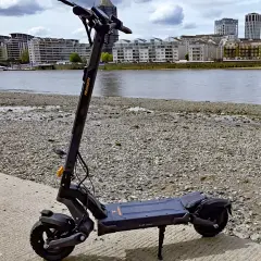 Trottinette électrique noire posée sur un chemin de gravier, avec une rivière et des immeubles en arrière-plan. Idéal pour savoir où garer sa trottinette électrique.