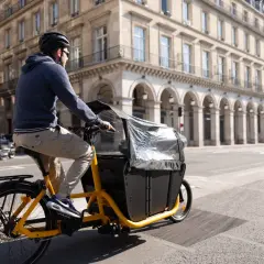 Un homme roule à Paris sur un vélo électrique cargo, prêt à transporter une charge lourde dans sa caisse noire.