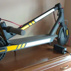 Trottinette électrique noire avec des bandes jaunes et blanches, des stickers réfléchissants pour plus de sécurité. Elle est branchée à son chargeur.