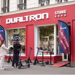 Devant le magasin DUALTRON STORE, un employé remet un casque à une cliente. Des trottinettes électriques sont exposées.