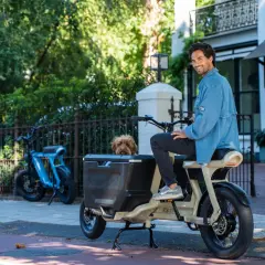 Un homme souriant est assis sur un fatbike cargo, son chien regardant depuis le panier. Un autre vélo bleu est garé à proximité.