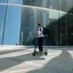 Livreur sur trottinette avec une grande sacoche noire, se déplaçant devant un immeuble moderne aux vitres bleues.