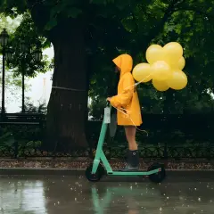 Une personne en manteau jaune conduit une trottinette ipx5 sous la pluie, tenant des ballons jaunes.