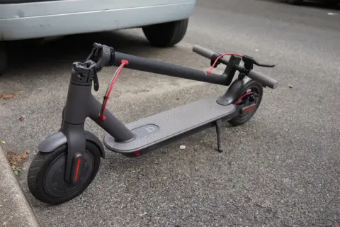Trottinette électrique grise, avec sa potence repliée, garée sur le bitume à côté d'une voiture.
