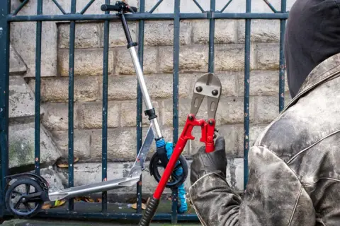 Un individu masqué, ganté, s'apprête à couper le cadenas d'une trottinette électrique avec une pince monstre.