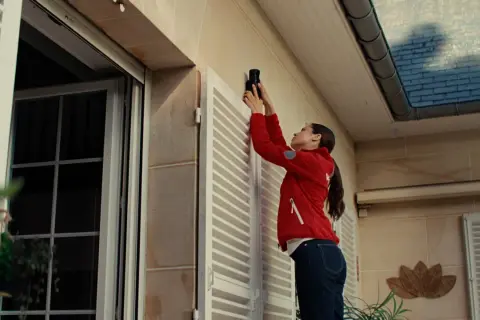 Une femme installe un appareil, peut-être une xiaomi alarme, sur le mur extérieur d'une maison, près d'une fenêtre.