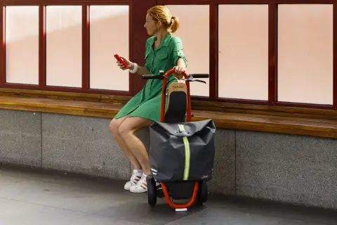 Femme en robe verte assise, tenant un téléphone rouge, à côté d'une trolley trottinette grise et orange.