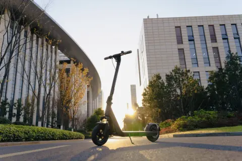 Trottinette électrique Kuickwheel S1-C Pro garée sur une route, avec des bâtiments modernes et des arbres en arrière-plan, au soleil couchant.