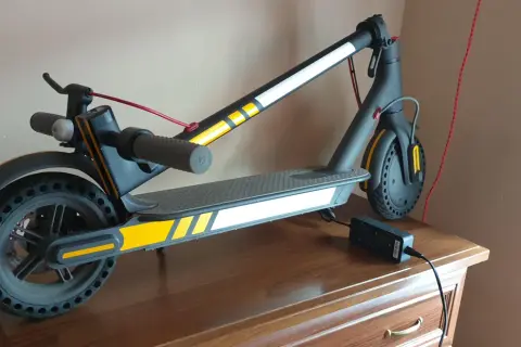 Trottinette électrique noire avec des bandes jaunes et blanches, des stickers réfléchissants pour plus de sécurité. Elle est branchée à son chargeur.