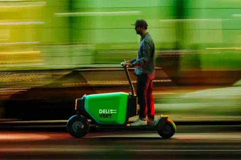 Un livreur sur une trottinette cargo verte "DELIVERY" file dans la nuit, le paysage flou derrière lui.