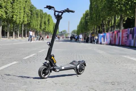 Trottinette électrique Dualtron Togo Plus sur une avenue bordée d'arbres, avec des passants et des décorations colorées en arrière-plan.