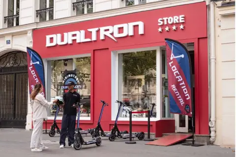 Devant le magasin DUALTRON STORE, un employé remet un casque à une cliente. Des trottinettes électriques sont exposées.