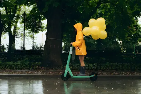 Une personne en manteau jaune conduit une trottinette ipx5 sous la pluie, tenant des ballons jaunes.