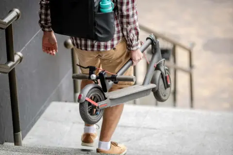Un homme porte sa trottinette électrique pliée, peut-être pour la vendre ou la transporter.