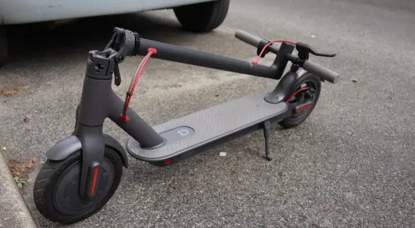 Trottinette électrique grise, avec sa potence repliée, garée sur le bitume à côté d'une voiture.