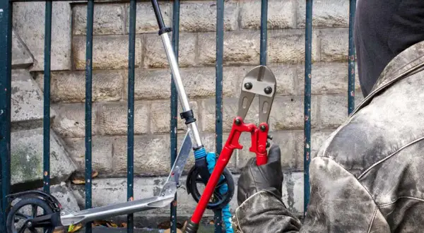 Un individu masqué, ganté, s'apprête à couper le cadenas d'une trottinette électrique avec une pince monstre.