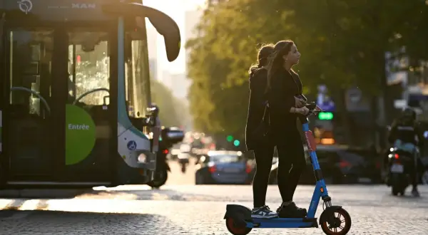 Deux jeunes femmes attendent sur une trottinette électrique, baignées par la lumière du soleil couchant. Un bus RATP est garé à gauche.