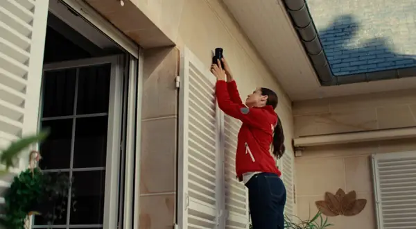 Une femme installe un appareil, peut-être une xiaomi alarme, sur le mur extérieur d'une maison, près d'une fenêtre.
