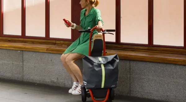 Femme en robe verte assise, tenant un téléphone rouge, à côté d'une trolley trottinette grise et orange.