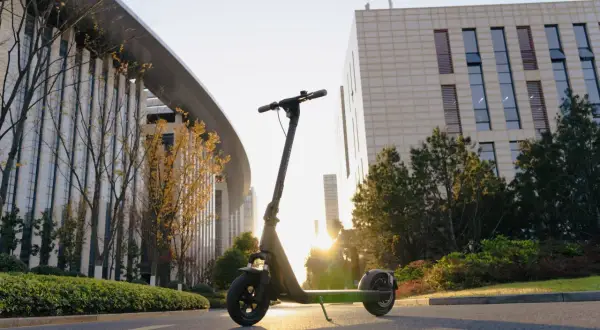 Trottinette électrique Kuickwheel S1-C Pro garée sur une route, avec des bâtiments modernes et des arbres en arrière-plan, au soleil couchant.