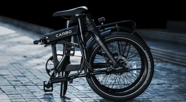 Un vélo électrique carbone pliant, noir, sur un pavé mouillé. Son design moderne et épuré promet des trajets urbains fluides.