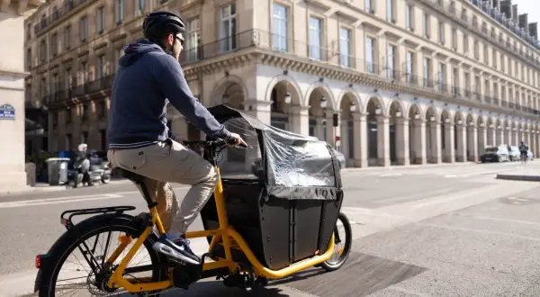 Un homme roule à Paris sur un vélo électrique cargo, prêt à transporter une charge lourde dans sa caisse noire.