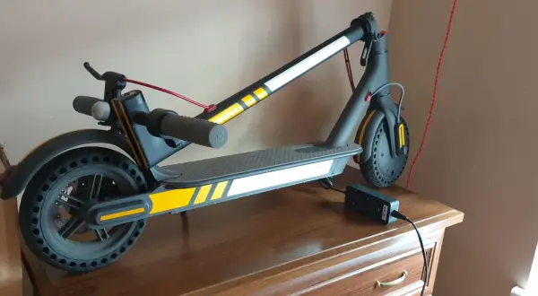 Trottinette électrique noire avec des bandes jaunes et blanches, des stickers réfléchissants pour plus de sécurité. Elle est branchée à son chargeur.