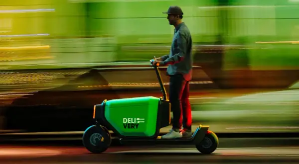 Un livreur sur une trottinette cargo verte "DELIVERY" file dans la nuit, le paysage flou derrière lui.