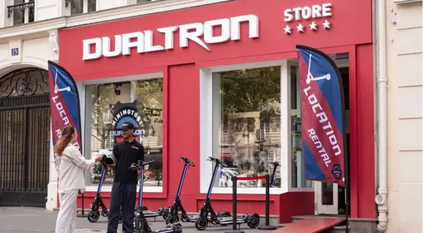 Devant le magasin DUALTRON STORE, un employé remet un casque à une cliente. Des trottinettes électriques sont exposées.