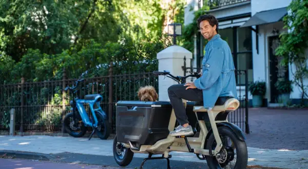 Un homme souriant est assis sur un fatbike cargo, son chien regardant depuis le panier. Un autre vélo bleu est garé à proximité.