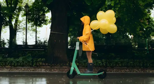 Une personne en manteau jaune conduit une trottinette ipx5 sous la pluie, tenant des ballons jaunes.