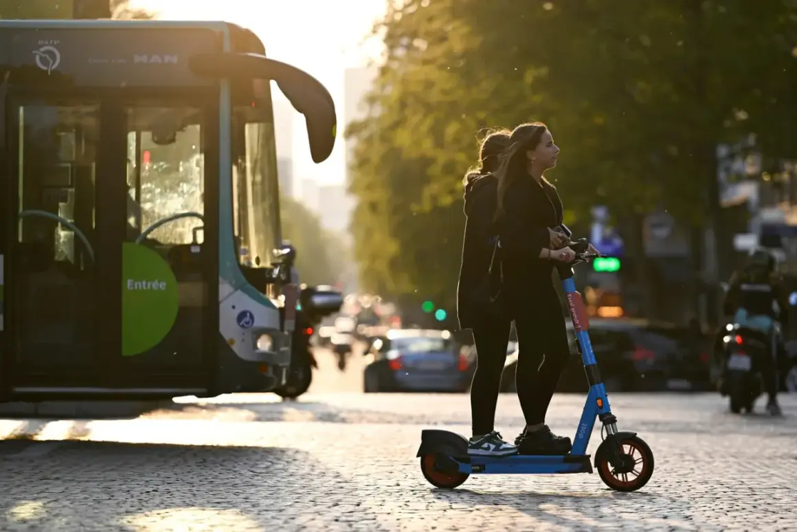 Deux jeunes femmes attendent sur une trottinette électrique, baignées par la lumière du soleil couchant. Un bus RATP est garé à gauche.