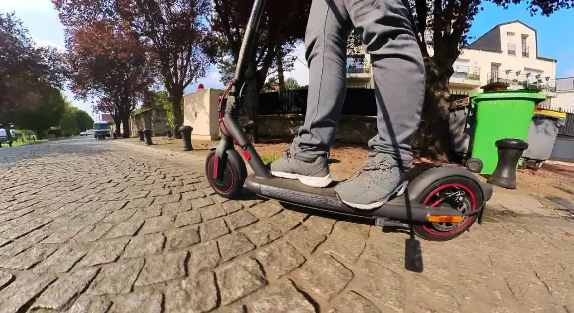 Pieds sur un trottinette pro 4 Xiaomi, roulant sur une rue pavée bordée d'arbres et de maisons.