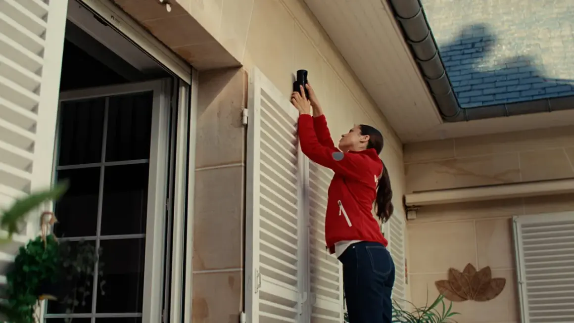Une femme installe un appareil, peut-être une xiaomi alarme, sur le mur extérieur d'une maison, près d'une fenêtre.