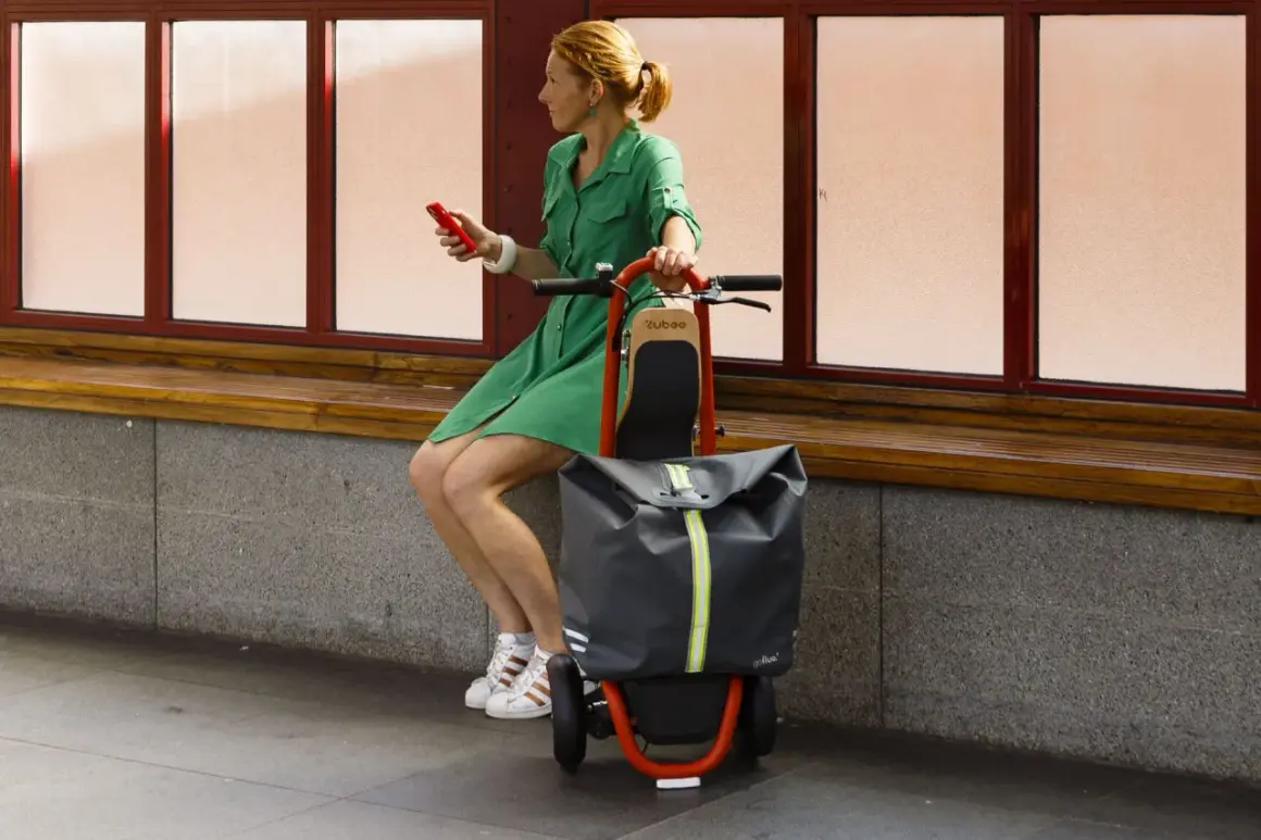Femme en robe verte assise, tenant un téléphone rouge, à côté d'une trolley trottinette grise et orange.
