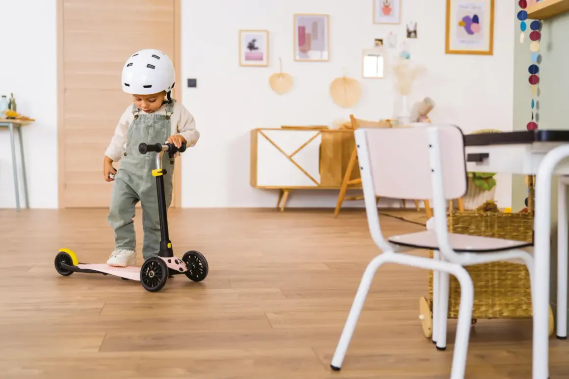 Un enfant porte un casque et se tient sur une trottinette rose. Il semble prêt à explorer, peut-être avec des stickers trottinette personnalisés pour ajouter une touche de couleur.