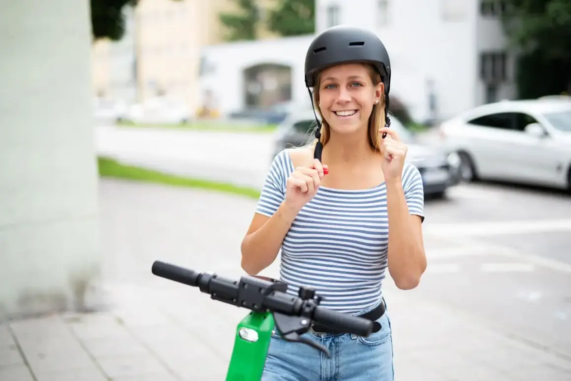 Jeune femme souriante ajustant son casque trottinette électrique, prête pour une balade en ville.