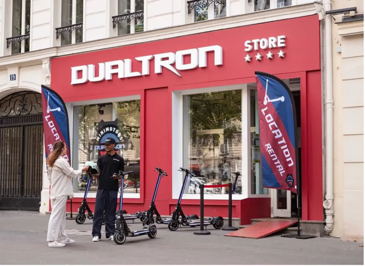 Devant le magasin DUALTRON STORE, un employé remet un casque à une cliente. Des trottinettes électriques sont exposées.