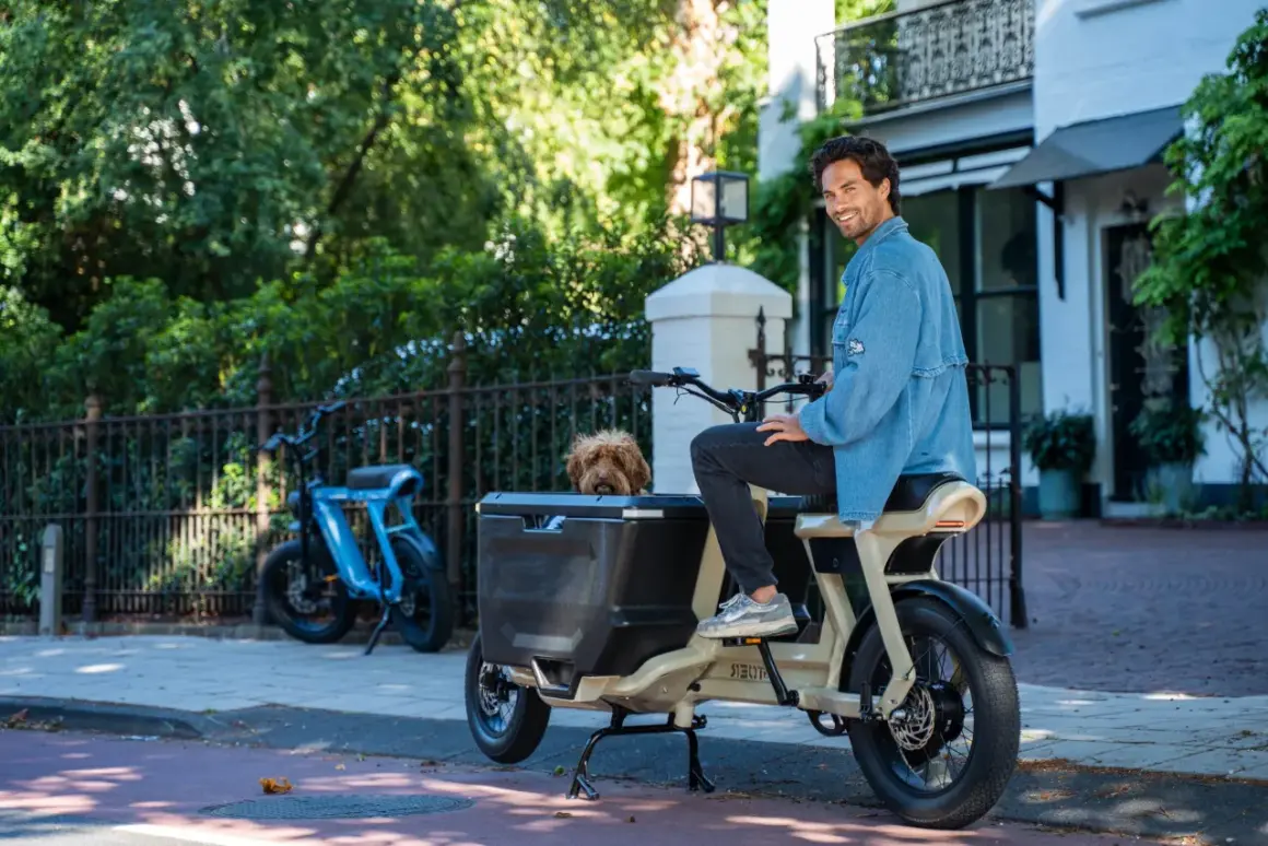 Un homme souriant est assis sur un fatbike cargo, son chien regardant depuis le panier. Un autre vélo bleu est garé à proximité.