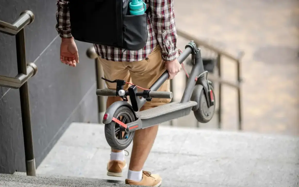 Un homme porte sa trottinette électrique pliée, peut-être pour la vendre ou la transporter.