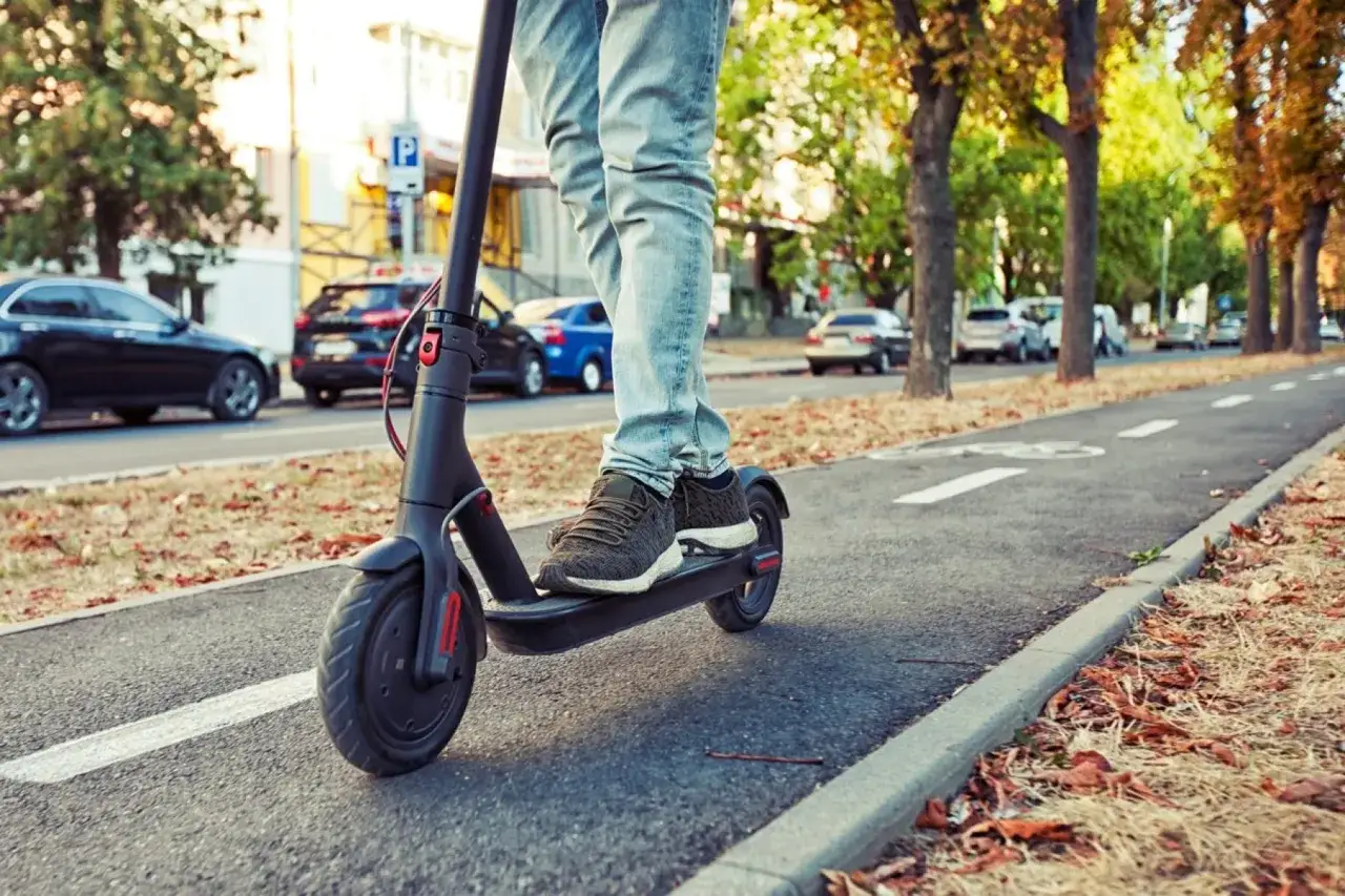 Un homme roule en trottinette électrique Xiaomi M365 sur une piste cyclable. Les feuilles d'automne jonchent le sol.