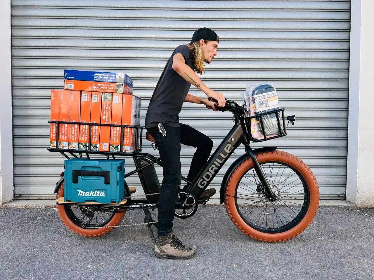 Un homme livre des colis sur un fatbike cargo. Il transporte des boîtes de porte-bagages et une caisse Makita.