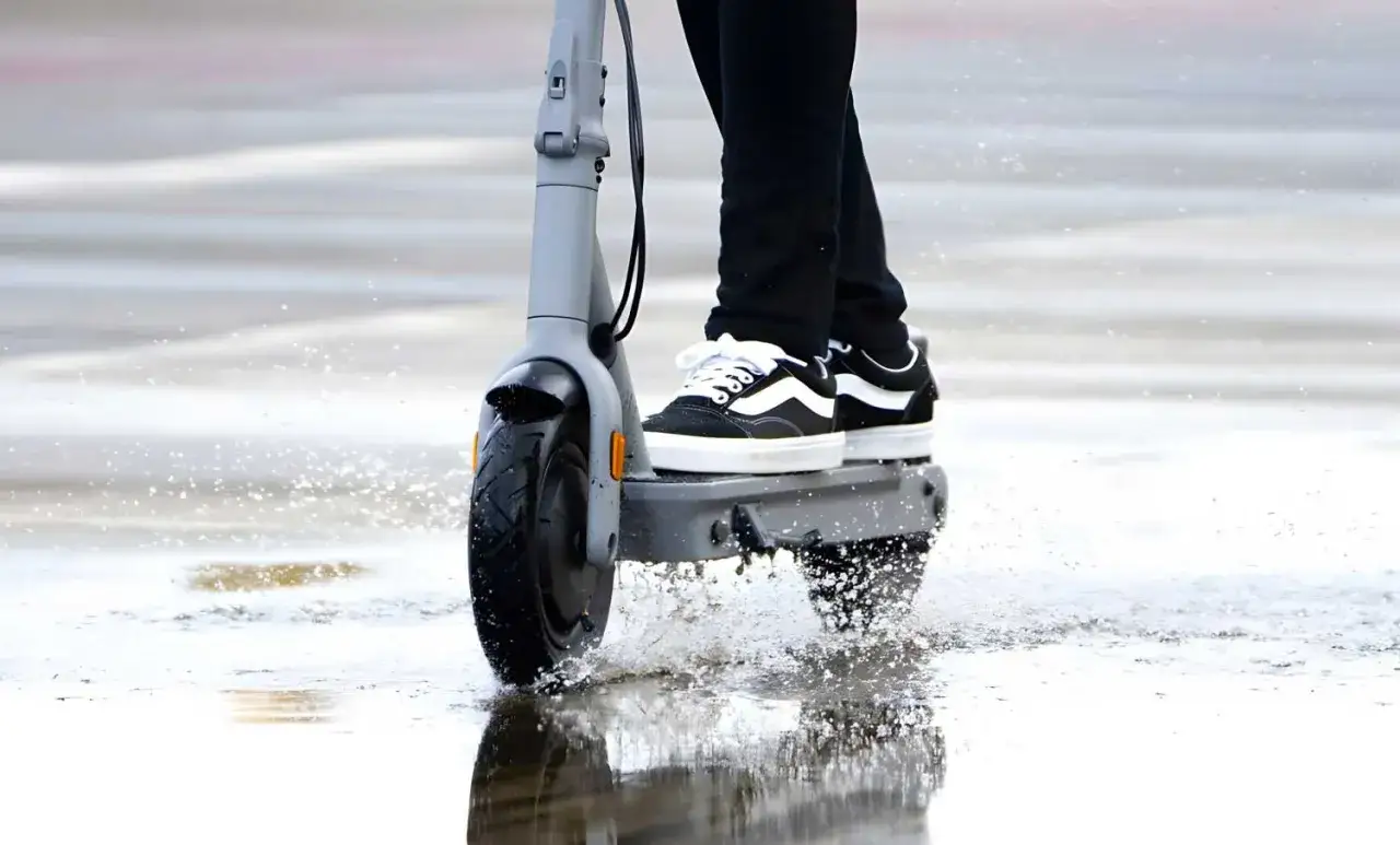 Pieds en baskets sur une trottinette, traversant des flaques d'eau. La protection pluie trottinette est essentielle pour rester au sec.