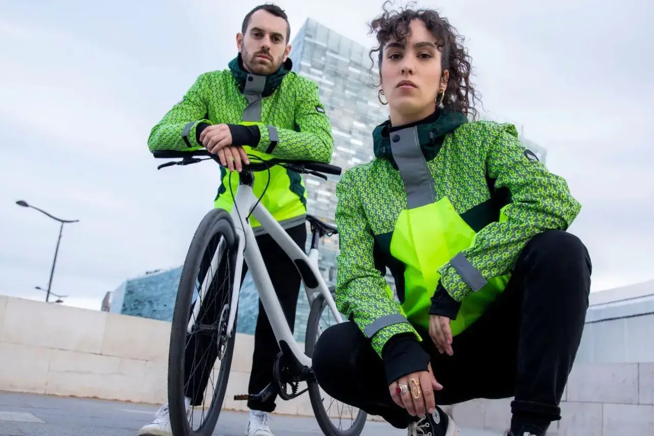 Deux cyclistes urbains portent des vestes fluo avec un motif discret. Leurs vêtements intègrent un dispositif réfléchissant pour une visibilité accrue.