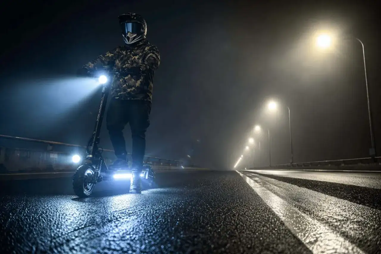 Un cycliste en casque et veste camouflage est sur une trottinette &eacute;lectrique, phares allum&eacute;s, sur une route mouill&eacute;e et brumeuse la nuit. Le clignotant trottinette est bien visible.