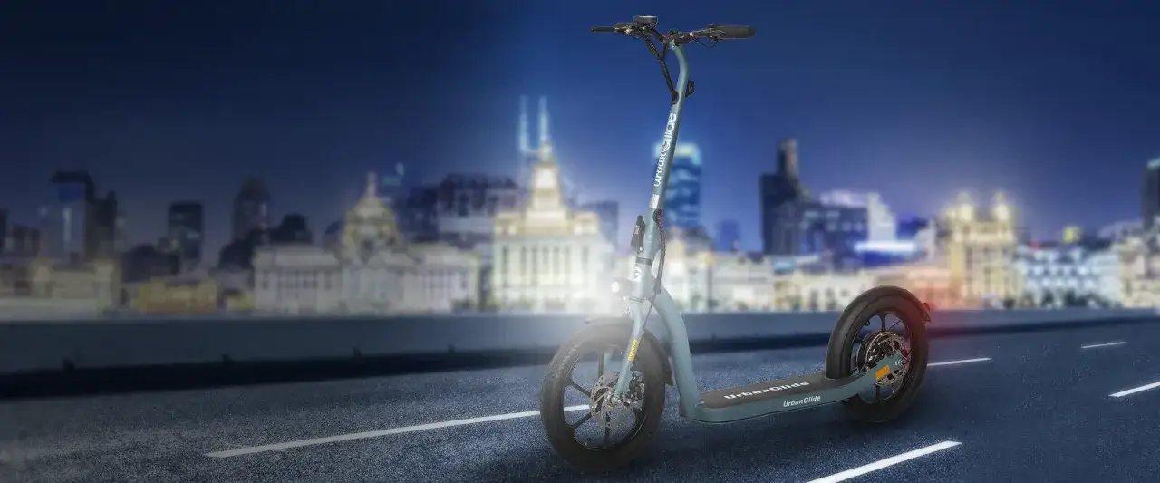Trottinette électrique UrbanGlide sur une route de nuit, avec une ville illuminée en arrière-plan. La lumière avant est allumée, prête pour l'aventure urbaine.